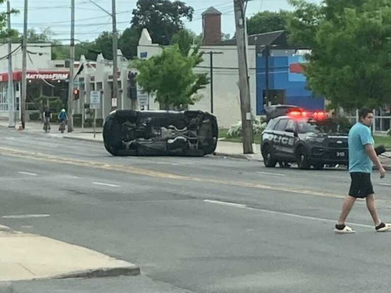 A car on its side in the middle of Boston Post Road near Bank of America, with a police car behind it and two cyclists nearby.