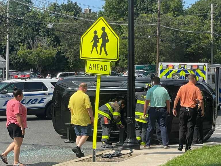 An SUV on its side on Boston Post Road beside a yellow school-crossing sign, with firefighters, police and an ambulance on the scene.