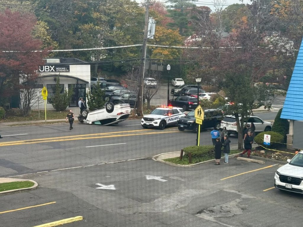 A white car upside down on Boston Post Road by IHOP, with police cars, officers and bystanders alongside two school-crossing signs.
