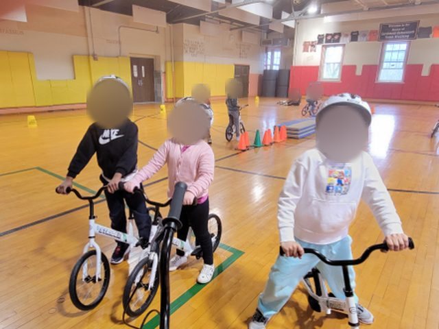 Kindergartners on balance bikes in a school gym, wearing helmets.