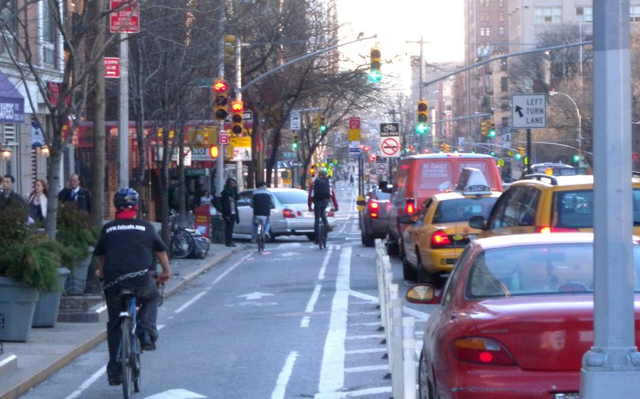 A cyclist in the protected bike lane on 9th Avenue in Manhattan, with a row of parked cars and traffic beside it.